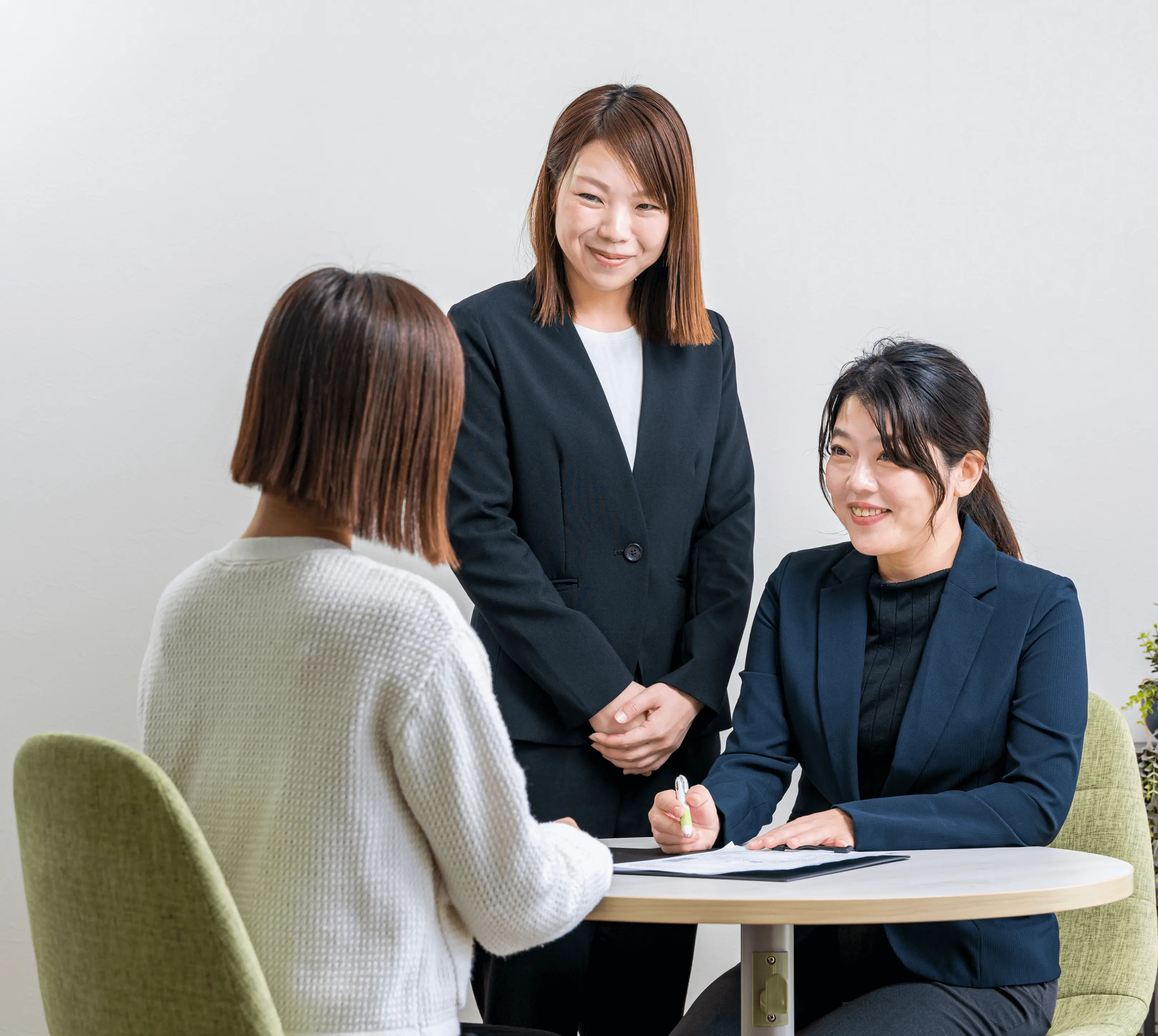 面談ブースでスタッフ2名が来訪者を笑顔で迎え、書類を確認しながら丁寧に説明している様子。女性スタッフが着席してペンを持ち、もう一人のスタッフが立ってサポートする姿から、安心感のある接客対応を伝えるビジネスシーン。