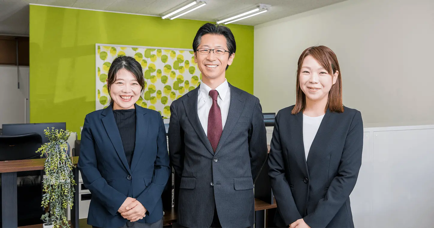 オフィスで笑顔のスタッフ3名が並んで立っている集合写真。明るい雰囲気の職場で、賃貸物件や住まい相談を丁寧にサポートする会社のスタッフ紹介をイメージした画像。